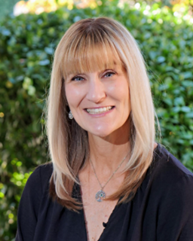 Smiling person with long, wavy blonde hair wearing a light blue scarf and black top against a dark background.